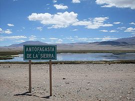 Vista de un cartel de ruta que anuncia Antofagasta de la Sierra: detrás una laguna y montañas
